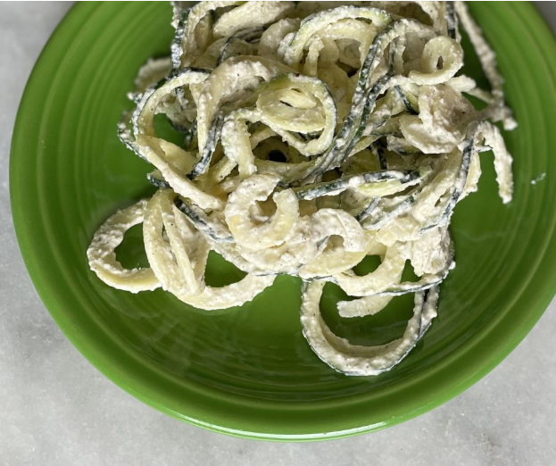 A green plate holds a serving of spiralized zucchini noodles coated in a creamy white sauce, arranged in a casual pile on a light-colored surface.