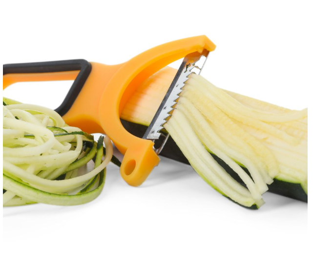 A close-up of a yellow vegetable peeler slicing a zucchini into thin, noodle-like strips, with some zucchini noodles piled beside the tool on a white background.