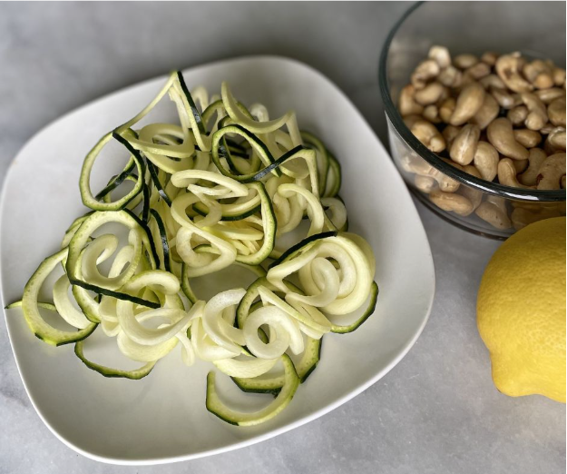 A plate of raw spiralized zucchini sits beside a glass bowl of cashews and a whole lemon on a gray surface.