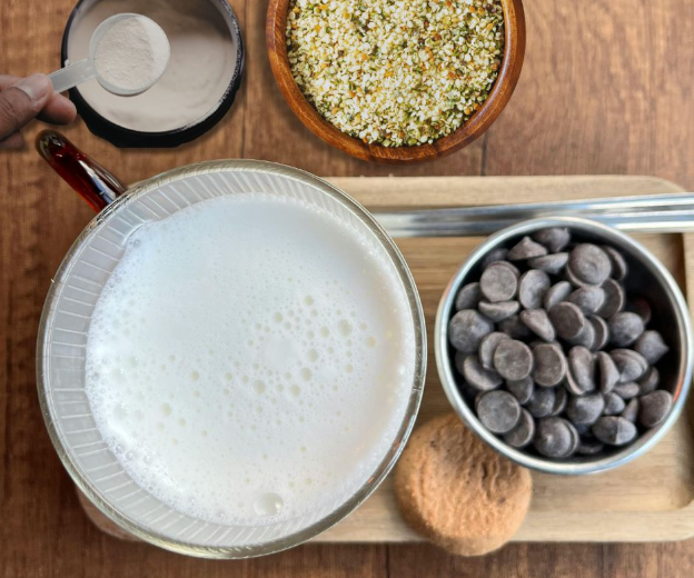 A top-down view of a cup of frothy milk, a hand holding a spoonful of powder, a bowl of hemp seeds, a bowl of chocolate chips, a cookie, and two metal straws on a wooden board.
