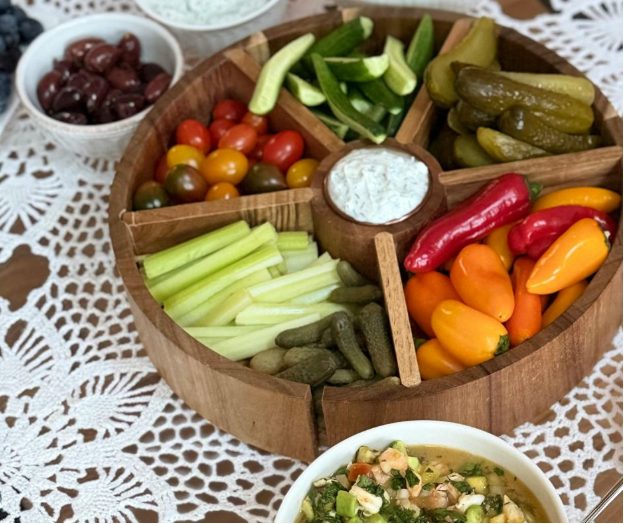 A wooden tray with sections filled with mini cucumbers, cherry tomatoes, pickles, celery sticks, colorful mini bell peppers, and a central dish of dip, set on a lace tablecloth. Olives and a bowl of salad are nearby.
