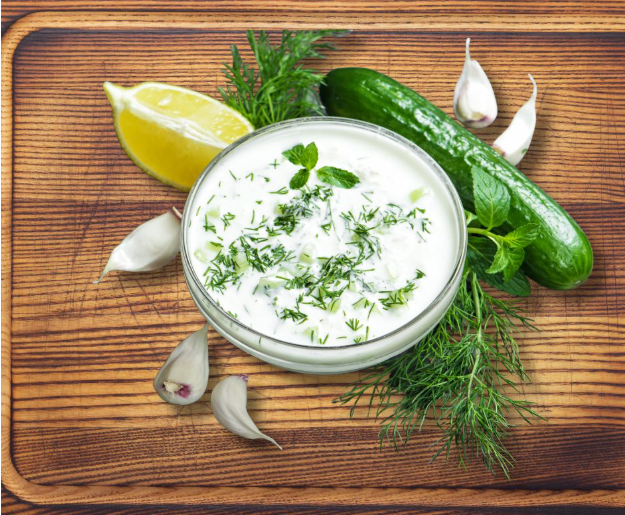 A bowl of tzatziki sauce garnished with fresh herbs, surrounded by a lemon wedge, garlic cloves, fresh dill, mint, and a cucumber on a wooden cutting board.