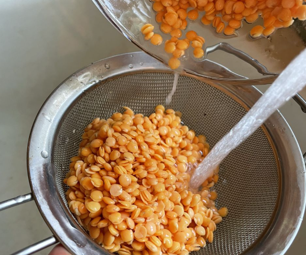 Red lentils being rinsed under running water in a metal strainer, with more lentils being poured in from a silver bowl above.