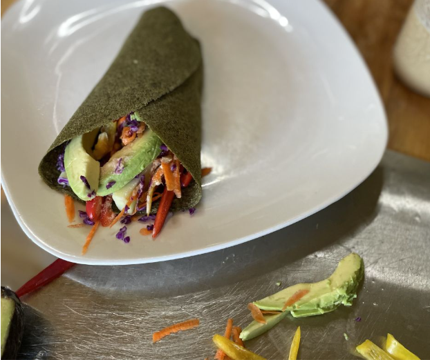 A green wrap filled with sliced avocado, shredded carrots, purple cabbage, and other vegetables sits on a white plate. Nearby, avocado slices and veggie strips are on a metal surface.
