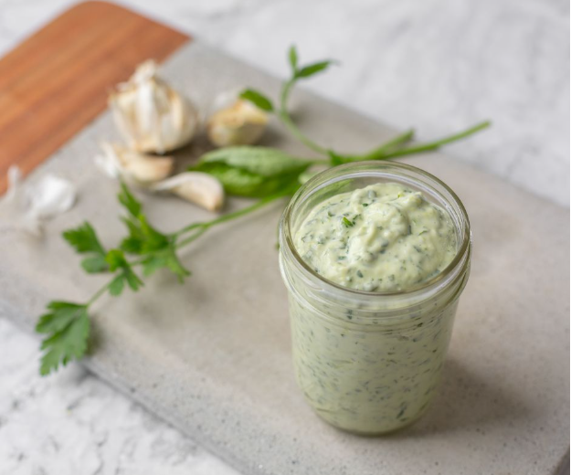 A jar of creamy green herb sauce sits on a gray tray, with garlic cloves and fresh parsley beside it.