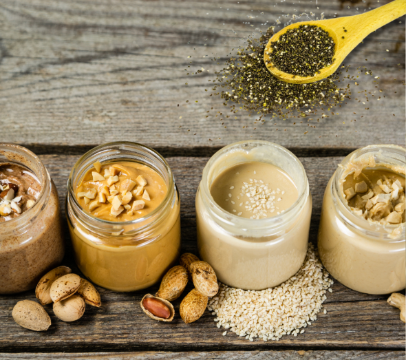 Four jars of nut and seed butters sit on a wooden surface, each topped with chopped nuts or seeds. Whole nuts and seeds are in front, with a spoon sprinkling chia seeds above.